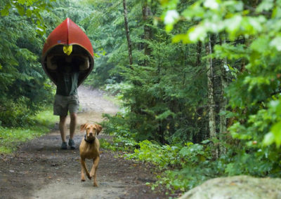 Story-telling the outdoor travel experience with pictures: A canoeist on the wooded Northern Forest Canoe Trail carries the red canoe through portage, with his dog running besides him, in the Adirondacks near Lake Placid, New York. Photo by Suzy Allman