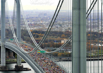 Crowded bridge at the start of the New York City marathon, showing packed lanes along the car-less bridge, and the neighborhoods of Staten Island in the background.