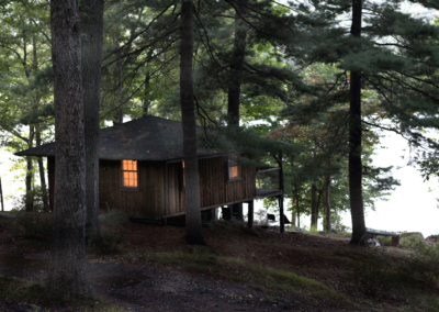 Baker Camp shuts down in the late fall after a summer of outdoor tourism. A cabin is perched in the trees at nightfall, with a single light on in the window, on the shores of Sebago Lake in Harriman State Park.
