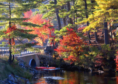 A red kayak slips through a culvert in Harriman State Park, surrounded by colorful trees of autumn and usually crowded with outdoor tourism.
