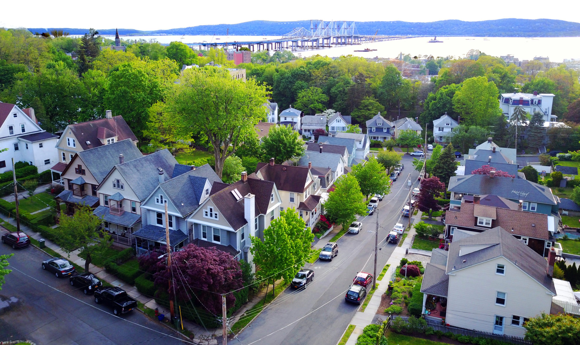 Overhead aerial view of neighborhood streets in Tarrytown, New York, showing rooftops, streets and the distant Hudson River. Recent work by Suzy Allman.