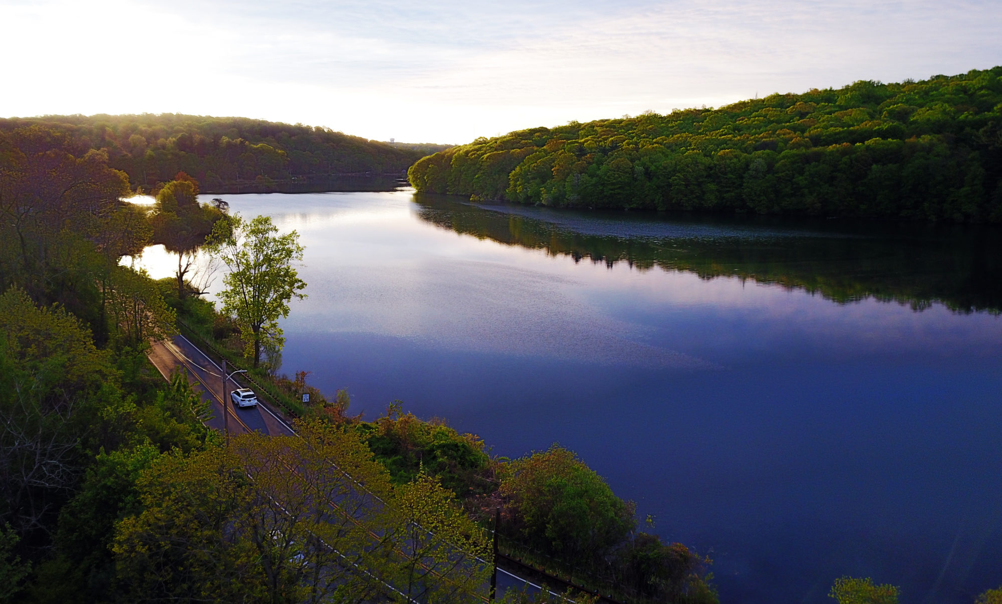 An overhead view of the Tarrytown Lakes, in Tarrytown New York, showing a single car on the roadway, a placid lake and rising sun.